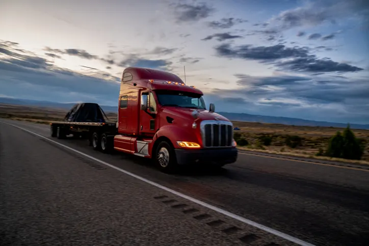 Trailer repair: flatbed, dry van, reefer, and lowboy service — Bledsoe OKC Red Peterbilt tractor pulling a loaded flatbed trailer on an Oklahoma highway — Bledsoe Truck & Trailer Repair services flatbeds, dry vans, reefers, and lowboys in the OKC metro.